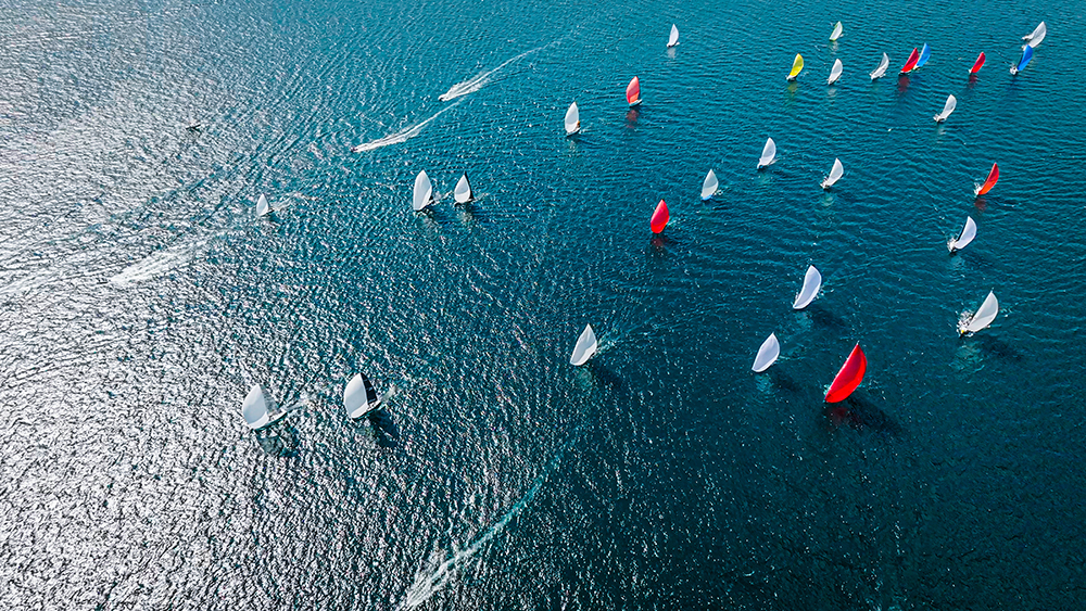 Aerial view of a wide sea with numerous sailboats scattered across the water, some with red and white sails billowing. The sunlight glistens on the rippling waves, creating a sparkling effect.