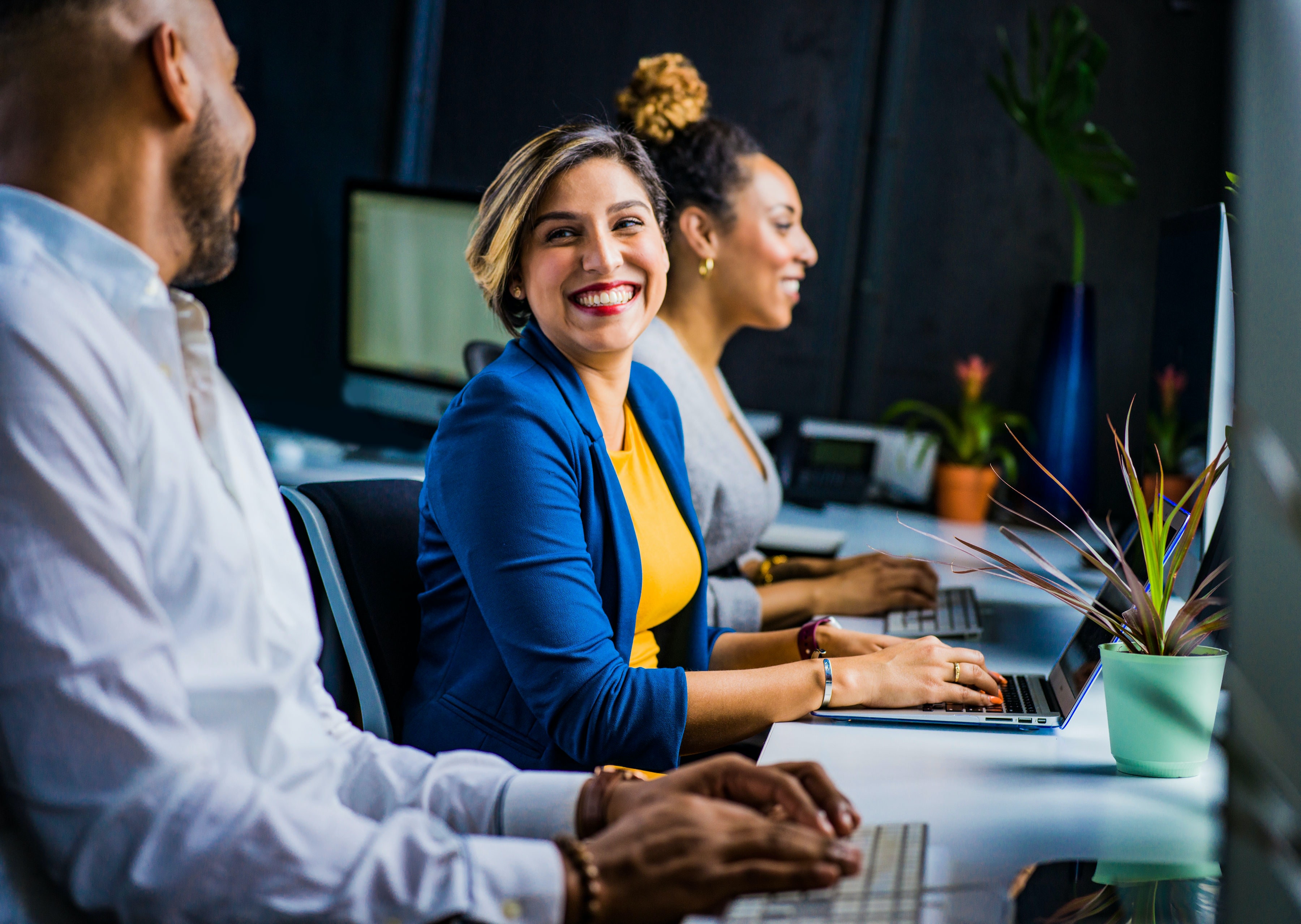woman in blue blazer and yellow top working at her computer with colleagues