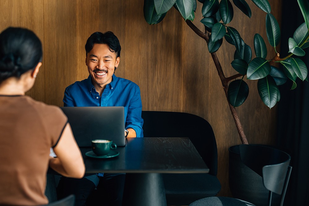 Asian man with mustache, in blue shirt, working at a coffee shop next to a plant