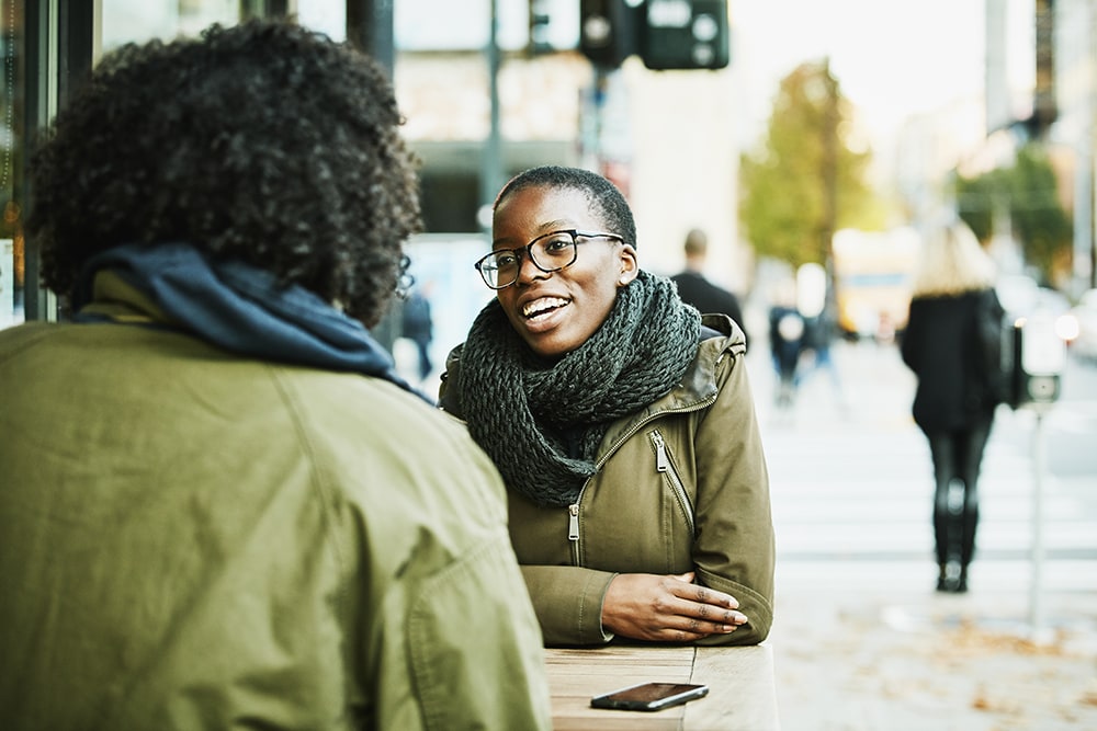 black person in green jacket chatting at outside cafe table with another person
