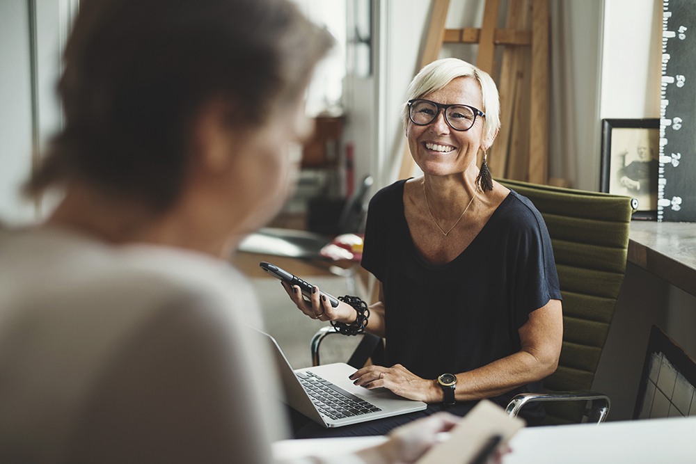 Woman smiling at man on her laptop with phone in hand
