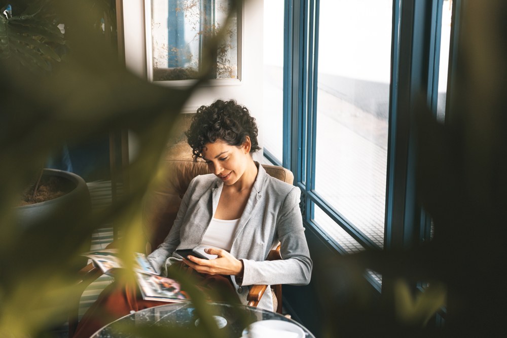 white woman with cropped brown hair on her phone at a cafe shown through blurred plant leaves