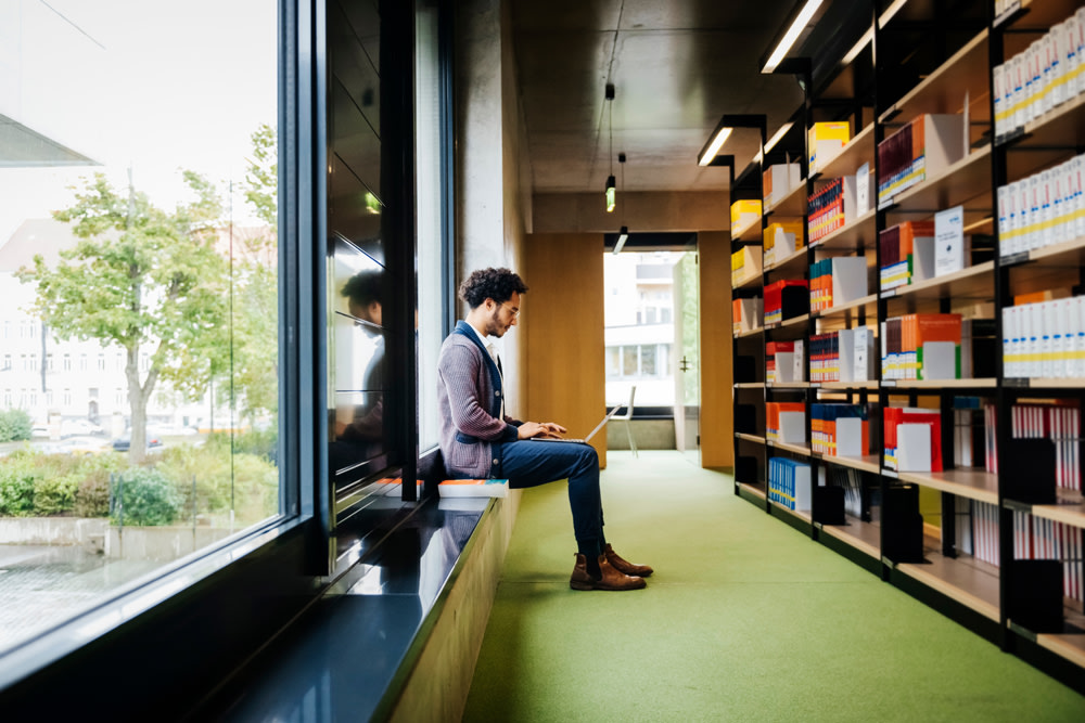 white man sitting with his laptop on the window sill in a library