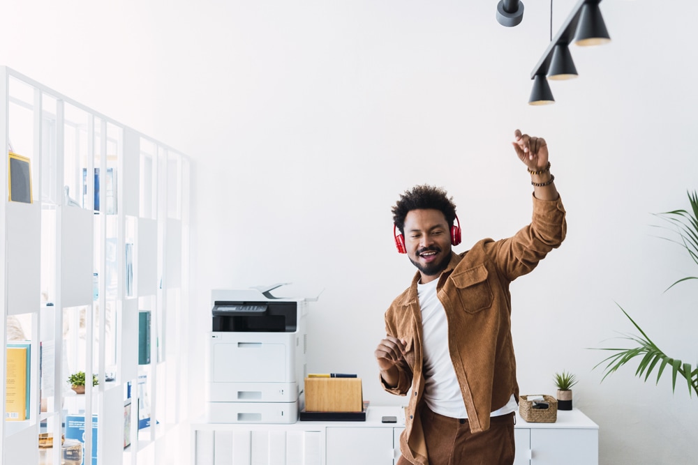 Black man in khaki jacket dancing in copy room with headphones 