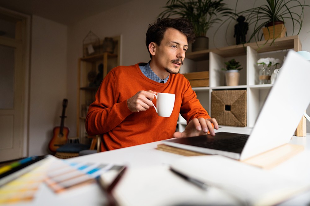 man with mustache in orange sweater drinking coffee at his computer