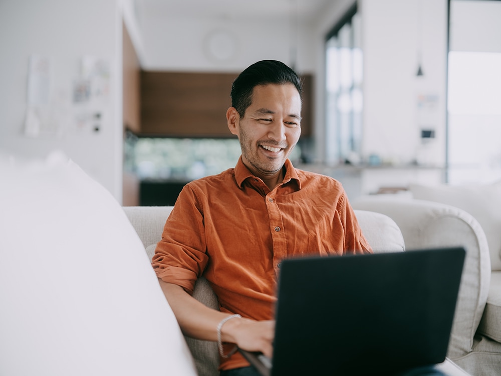 Asian man in orange shirt working at his laptop on his couch