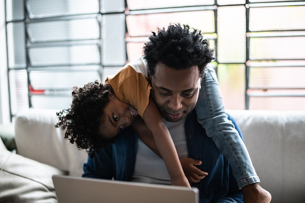 Black man on laptop with his son across his shoulders
