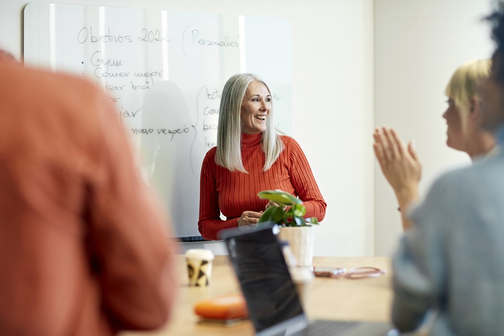 white woman with white hair in an orange sweater leading a discussion at work