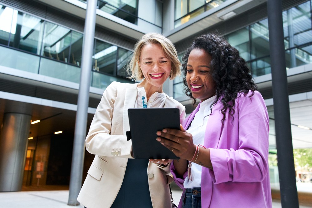 Two women in professional dress outside a corporate building happily looking at a tablet