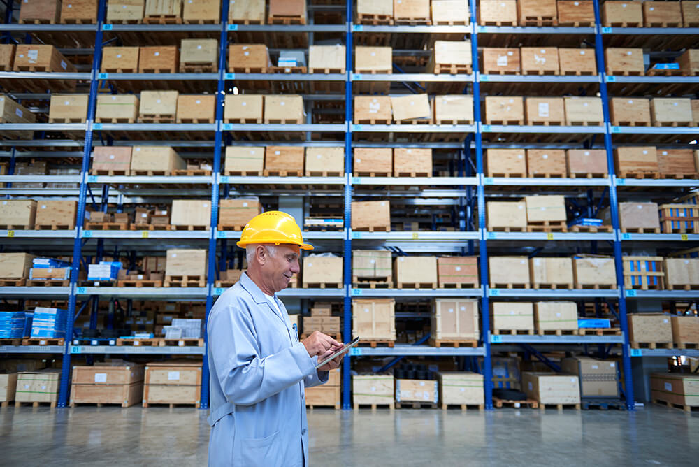 Middle aged white man with white hair and mustache wearing a yellow hard hat walking through a warehouse while working on a tablet