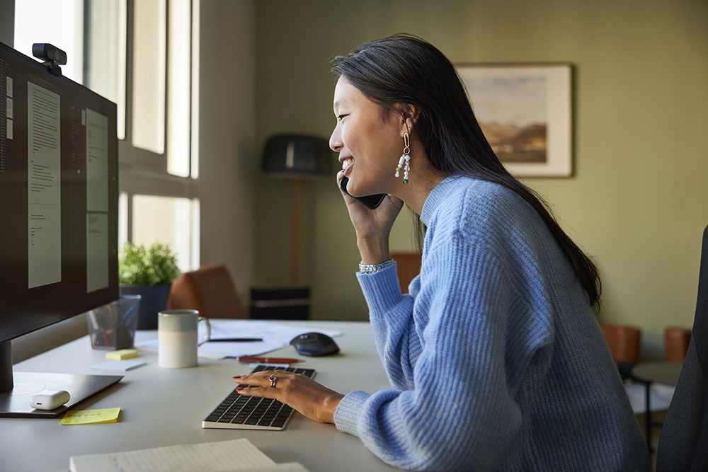 Asian woman in blue sweater talking on her phone at her computer