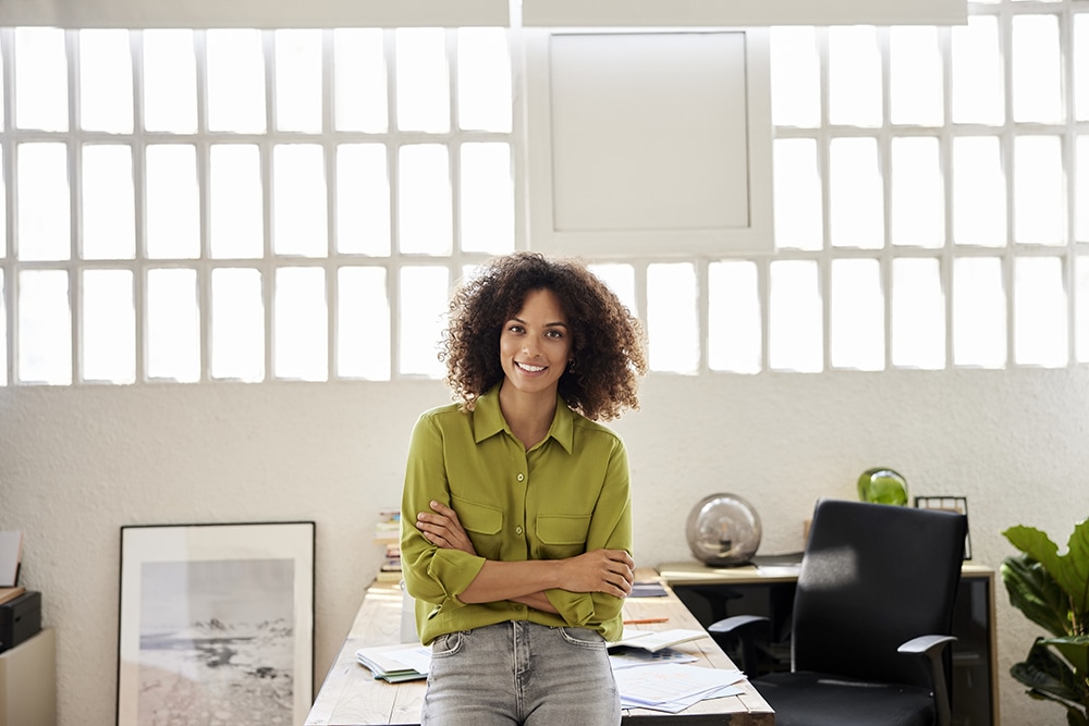 Black woman in green shirt and jeans leaning against her office desk